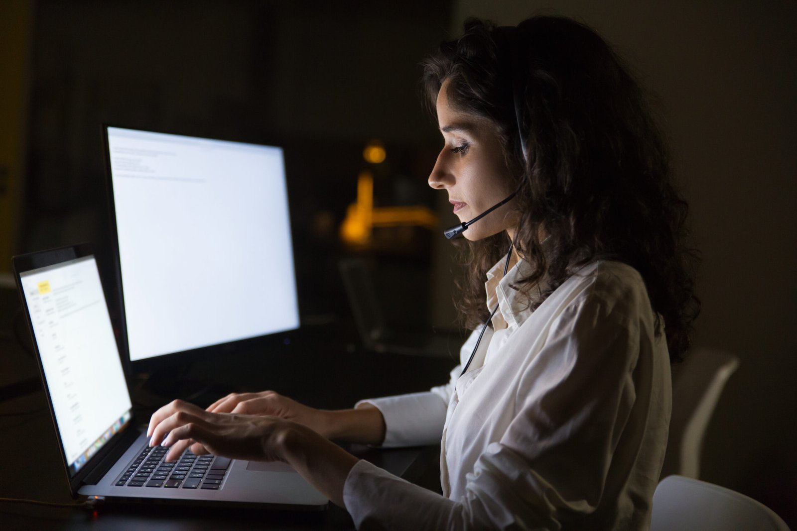 Focused businesswoman in headset using laptop. Side view of young serious call center operator working with laptop computer in dark office. Client service concept